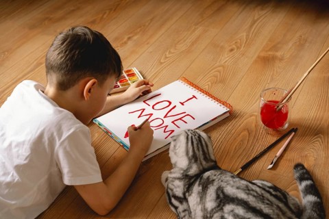 A young boy lies on the floor, diligently creating a heartfelt Mother's Day card, with the message