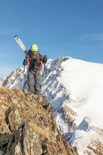 Skier in vibrant gear traverses rugged, snow-capped mountain terrain under a clear blue sky. The