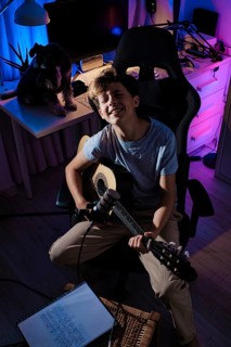 A young guitarist smiles while playing acoustic guitar in a cozy, vibrantly lit home studio with a