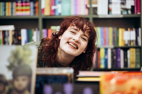 Portrait of a cheerful young woman with curly hair, smiling near a bookcase filled with colorful