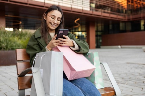 Smiling young woman sitting on a bench with shopping bags, looking at her phone She wears glasses