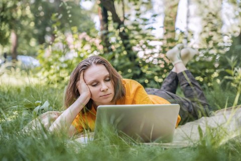 A woman lies on a blanket in a park, focused on her laptop. She enjoys the blend of technology and