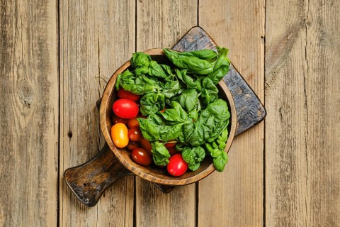 A wooden bowl holds fresh basil and cherry tomatoes on a rustic wooden table. The ingredients are