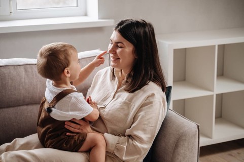 A smiling mother embraces her son, who playfully touches her nose. They are seated on a comfortable