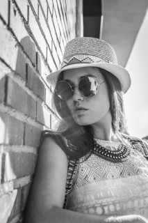A black and white photo of a woman in round sunglasses and a patterned hat, leaning casually
