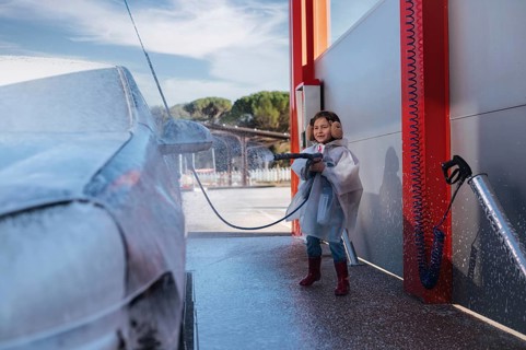 A smiling child in earmuffs and a raincoat playfully washes a car at a self service station,