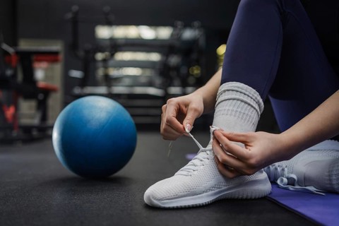 A close up of hands of woman tying white athletic shoes on a gym floor, with a blue exercise ball