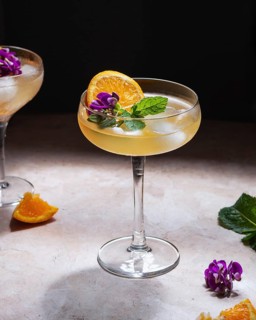 Cocktails in two glass is elegantly garnished with a slice of orange, vibrant purple flowers, and
