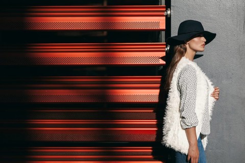 A stylish woman wearing a wide-brimmed hat and textured vest stands against a modern red and gray