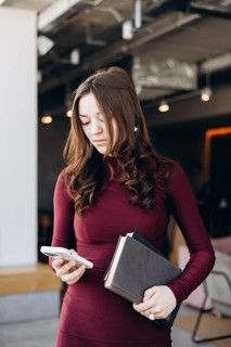 Female student in a maroon dress multitasking, holding books and using a smartphone, ready to study