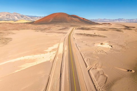 A long, straight road cuts through a vast desert, leading towards a distant, brown hued mountain