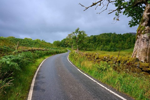 A country road crosses a green landscape flanked by trees and a stone wall under a cloudy sky,