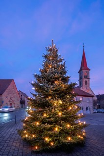 Illuminated Christmas tree on the village square at dusk, St Bartholomew's Church in the