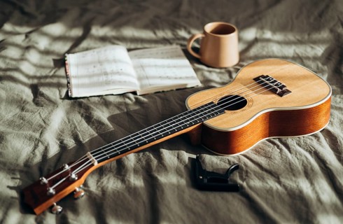 A ukulele rests on a cozy bed beside an open notebook and a warm mug, bathed in gentle natural