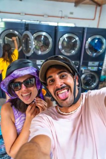 Happy couple wearing motorcycle helmets taking a selfie sticking out their tongues in a laundromat