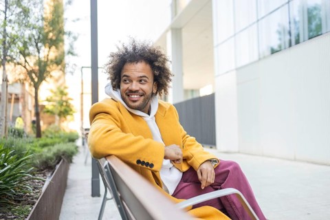 A cheerful afro man sits comfortably on a bench in a modern urban environment, wearing a stylish