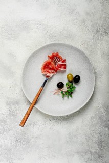 Prosciutto on a fork, top view, olives and micro-greens on a plate, light background, no people