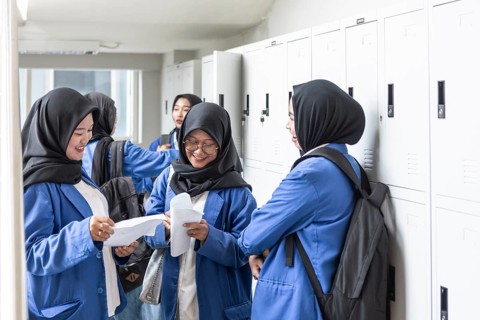 Asian Muslim students in blue uniforms happily share papers in a hallway, standing near lockers.