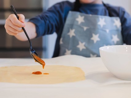A child prepares homemade pizza, spreading tomato sauce on a rolled out dough using a spoon. They
