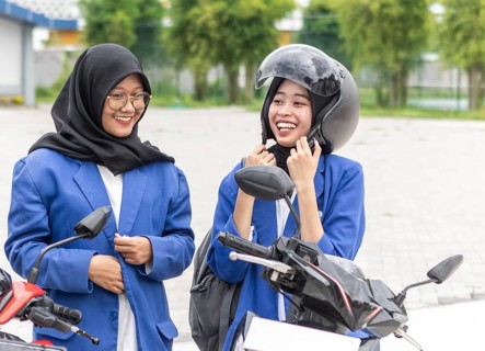 Two smiling Asian Muslim students in blue blazers stand beside their motorcycles, preparing for