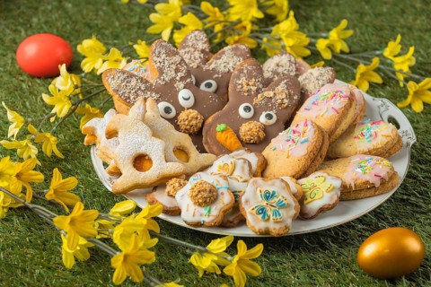 Various decorated Easter cookies on a plate next to flowers and colorful Easter eggs in the grass