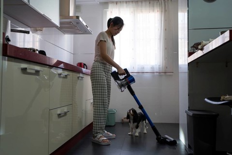 A woman in comfortable attire uses a cordless vacuum cleaner on a kitchen floor, with her pet dog