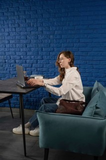 A woman in casual attire works on her laptop in a modern cafe with a vibrant blue brick wall. The