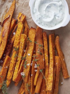 Crispy sweet potato fries on brown baking paper beside yogurt sauce bowl, close up view directly