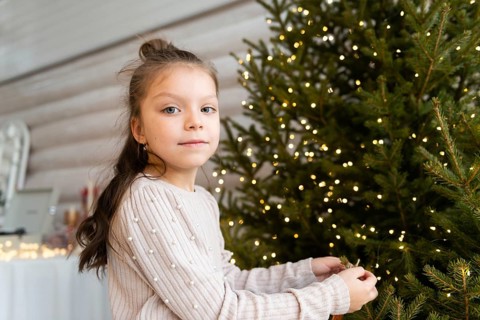 A young girl with a gentle expression is seen decorating a lush Christmas tree, which is brightly