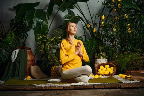 A woman sits on a mat in a garden filled with plants and lemons. She holds her hands together in a