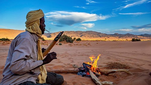 A man sits around a campfire in a vast desert landscape under a clear sky, Tuareg around a campfire