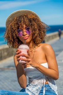 Vertical portrait of a young stylish latin woman drinking iced juice sitting next to the sea