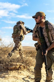 Two soldiers in camouflage gear navigate a sandy desert landscape, focused on a mission, with clear