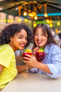 Vertical portrait of lesbian multi-ethnic couple enjoying cocktails smiling at camera in an outdoor