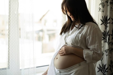 A serene pregnant woman stands by a sunlit window, gently holding her belly. She is immersed in
