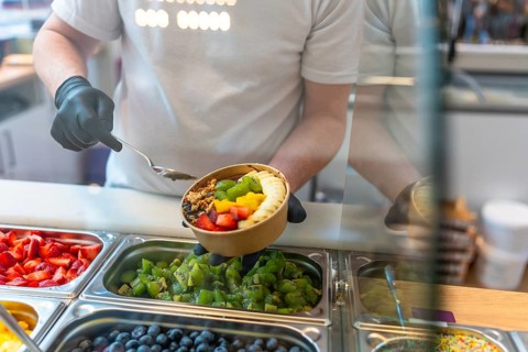 Person wearing black gloves holding a kraft paper bowl filled with acai, fresh strawberries, kiwi,