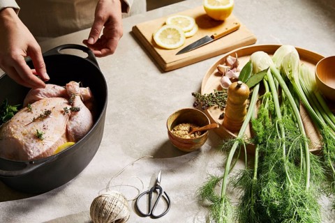 A cook seasons a whole chicken with fresh herbs and lemon in a kitchen setting. Fennel, garlic, and