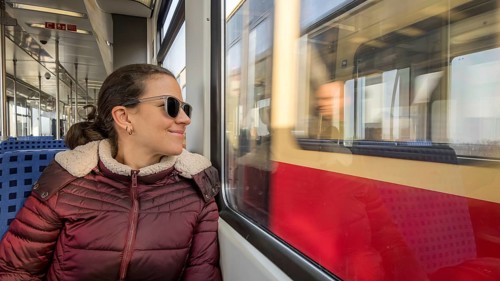A woman in sunglasses and a cozy jacket enjoys a scenic train ride on a sunny day, gazing out the
