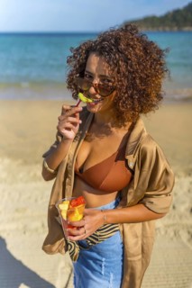 Vertical photo of a Confident arab woman biting a slice of tropical fruit on the beach