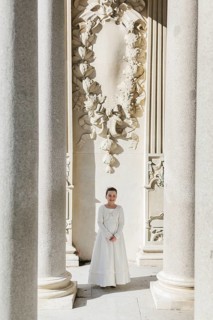 A young girl in a First Communion dress stands between majestic stone columns in a sunlit setting,