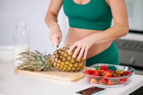 A pregnant woman in a green yoga outfit is cutting a fresh pineapple on a wooden board.