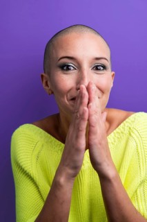 A middle aged woman with a shaved head is laughing and covering her mouth with her hands, dressed