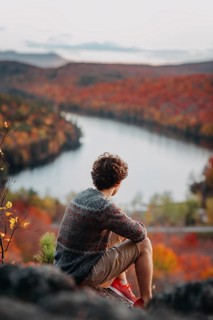 A man sits on a rocky ledge overlooking a river winding through a vibrant, colorful autumn
