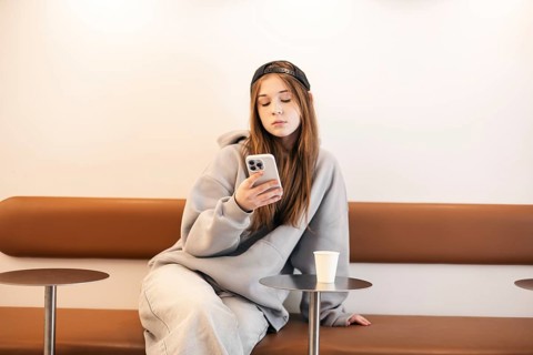 A teenage girl sitting on a stylish bench, wearing casual clothing and a cap, attentively using a