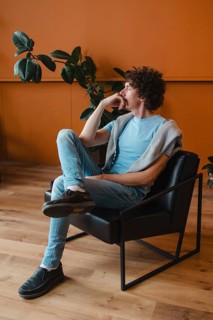 A young man with curly hair sits thoughtfully in a modern chair, wearing casual attire. Behind him,