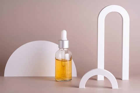 A cosmetic in glass dropper bottle filled with golden oil stands against a beige background,