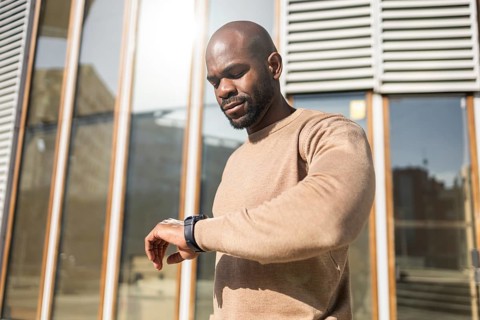 A cuban man in a brown sweater checks his watch outside on a sunny day. He stands near a modern