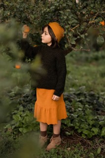 A girl wearing a cozy sweater and hat stands in a lush garden, reaching for an orange on a tree.