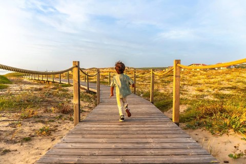 Young boy running on a wooden boardwalk leading through sand dunes towards the beach, experiencing