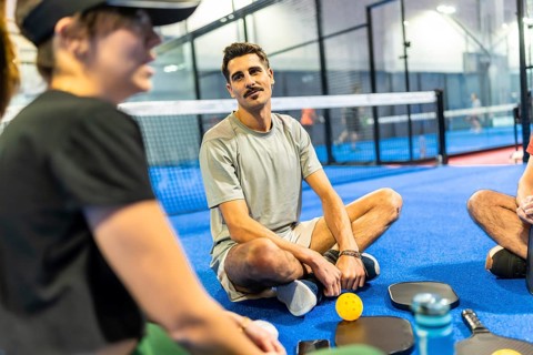 Friends resting and chatting on a blue indoor pickleball court after a game, enjoying conversation,